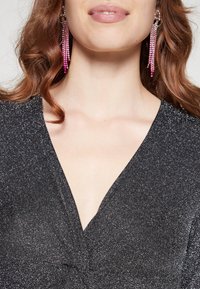 Svart klänning med V-ringning, tillverkad av ett glimrande material, med en vriden detalj vid midjan och trådar som hänger av rosa kristallörhängen.