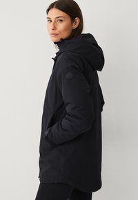 Vrouw in een zwarte winterjas met capuchon, met haar handen in de zakken, die over haar linkerschouder kijkt tegen een effen achtergrond.