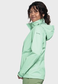 Giacca impermeabile verde chiaro con cappuccio, dotata di zip, tasche laterali e un design elegante. Logo del marchio visibile sulla manica.