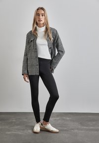 Veste chemise à motif pied-de-cochon noir et blanc sur un col roulé blanc, assortie de leggings noirs et de baskets beiges/blancs.
