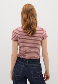 Persoon met schouderlang golvend rood haar, gekleed in een rood-wit gestreept T-shirt met korte mouwen en donkerblauwe jeans, met de rug naar de camera gekeerd.