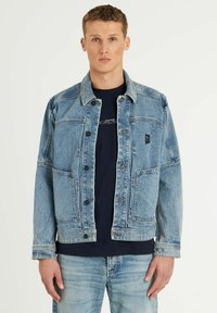 Chaqueta de mezclilla azul claro con un collar clásico, botones en la parte delantera y dos grandes bolsillos en el pecho. Combinada con una camisa azul marino debajo.