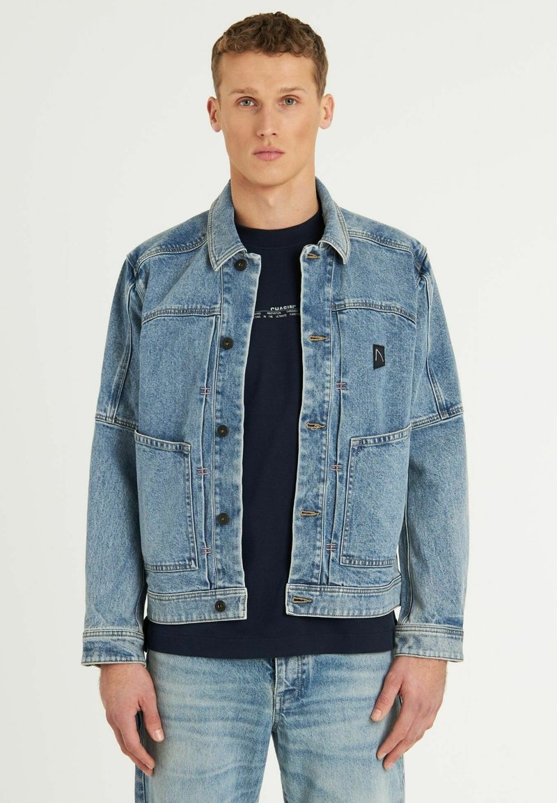 Chaqueta de mezclilla azul claro con un collar clásico, botones en la parte delantera y dos grandes bolsillos en el pecho. Combinada con una camisa azul marino debajo.