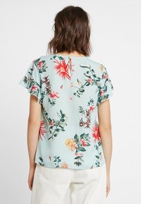 Květinová blůza světle modré barvy s volánkovými krátkými rukávy, zdobená červenými, růžovými a žlutými květy. Detaily s klíčovou dírkou na zadní straně. Hladký materiál.