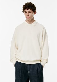 Pulover bej cu guler clasic, mâneci lungi și croială lejeră. Țesătură moale, design minimalist, asortat cu pantaloni din denim închis.