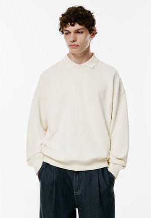 Beiger Pullover mit klassischem Kragen, langen Ärmeln und lockerer Passform. Weicher Stoff, minimalistisches Design, kombiniert mit dunklen Jeanshosen.