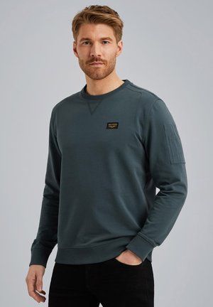 Mørk teal sweatshirt i et blødt, tekstureret stof. Har en ribbet krave, lange ærmer og en brystlappe med branddetaljer.