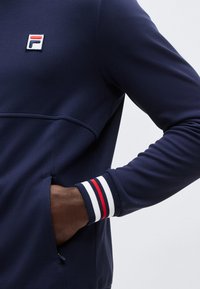 Χέρι με σκουρόχρωμο δέρμα σε τσέπη σακακιού σκούρου μπλε με ριγωτή μανσέτα; μικρό λογότυπο Fila στο στήθος.