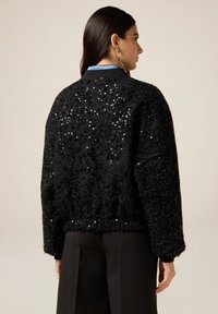 Giacca bomber nera con superficie testurizzata e paillettes sparse. Presenta un orlo e polsini a coste, con una vestibilità leggermente ampia.