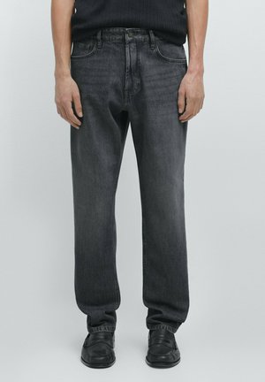 Muž v černých džínech rovného střihu, černých slip-on botách a černém žebrovaném svetr stojí na jednobarevném světlém pozadí.