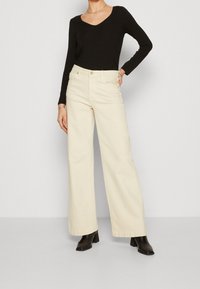 Pantaloni larghi a vita alta in tessuto misto cotone di colore avorio, abbinati a un top nero a maniche lunghe. Le scarpe sono stivaletti neri con tacco.