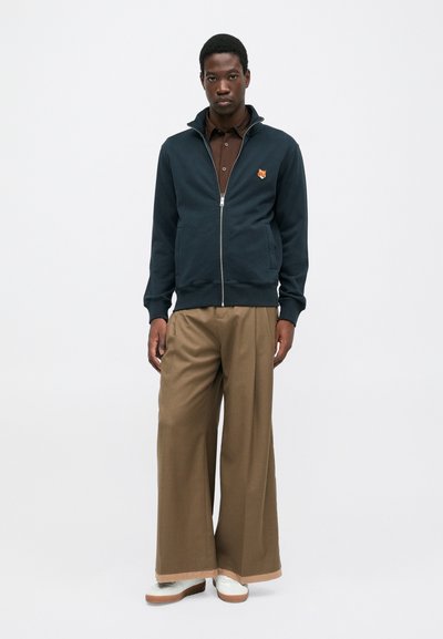 Felpa con zip blu navy e un piccolo logo di volpe arancione, abbinata a pantaloni larghi marroni e sneakers bianche, su uno sfondo neutro.