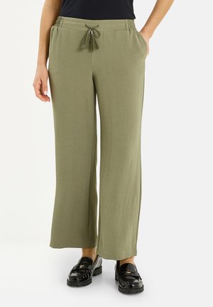 Mujer con pantalones anchos de pierna en verde oliva con cintura elástica y cordón con borlas, combinados con mocasines negros de charol.