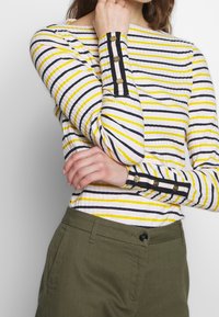 Randig långärmad topp med vita, gula och svarta linjer. Har knappramar på ärmsluten och ribbad textur. Kombineras med olivgröna byxor.