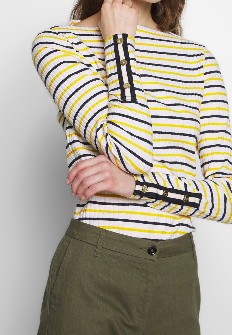 Randig långärmad topp med vita, gula och svarta linjer. Har knappramar på ärmsluten och ribbad textur. Kombineras med olivgröna byxor.