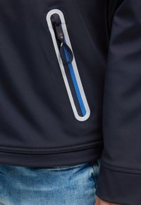 Chaqueta softshell de marina con detalle de bolsillo con cremallera en azul y gris, textura suave y costura visible a lo largo del dobladillo.