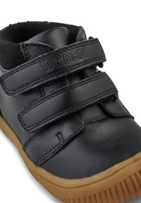 Chaussure enfant en cuir noir avec deux bandes velcro, de la marque "WODEN", et une semelle en caoutchouc marron clair.