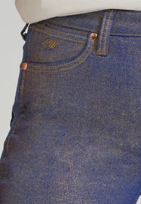 Plave traperice s zlatim šavovima, diskretnim logotipom na džepu i bakrenim detaljima. Glatka tekstura s blagim sjajem.