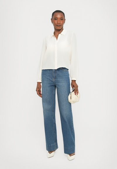 White, long-sleeve, button-up blouse with a scalloped hem, paired with light blue, wide-leg jeans and off-white slingback shoes, holding a cream clutch.