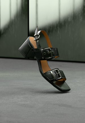 Sandales à talons en cuir noir avec des larges brides munies de boucles argentées ajustables, bout carré et talon block. Texture lisse, design minimaliste.