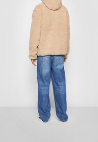 Giacca in pile beige con cappuccio, dal morbido aspetto tessurizzato, abbinata a jeans blu larghi con un lavaggio chiaro e tasche posteriori.