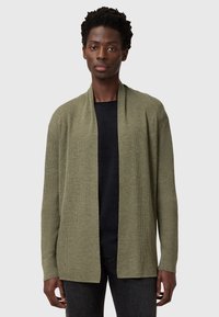 Olivgrüner, offener Cardigan mit gerippter Textur und langen Ärmeln, über einem schwarzen Shirt getragen, kombiniert mit dunklen Jeans.