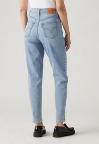 Person som bär ljusblå högmidjade Levi's jeans och svarta loafers, stående med ryggen mot en enfärgad bakgrund.