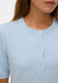 Donna che indossa un cardigan lavorato a maglia blu chiaro con maniche corte e bottoni bianchi a forma di cuore, che mostra il colletto e la parte superiore del torso.