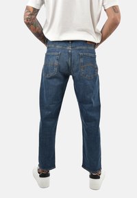 Jeans in denim blu scuro con taglio a gamba dritta, cinque tasche e dettagli di cuciture visibili, indossati con calzature bianche.