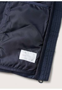 Marinblå kviltad jacka inuti med slät foder, som har en tygetikett för namn och telefonuppgifter, samt en dragkedjeficka.