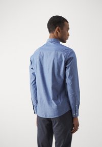Camisa azul de botones con un patrón sutil, mangas largas y un diseño en el cuello trasero, combinada con pantalones gris oscuro. Textura de tela suave.