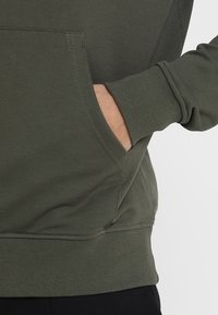 Sudadera de color verde oliva con un bolsillo canguro, puños y dobladillo acanalados. Fabricada en una suave mezcla de algodón con una textura lisa y detalles mínimos.