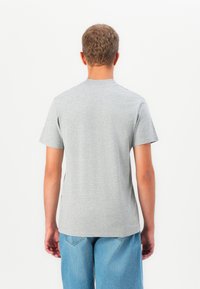 Uomo con capelli ricci corti che indossa una maglietta a maniche corte grigia chiara e jeans azzurri, voltato di spalle su uno sfondo bianco.
