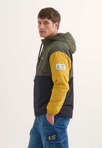 Chaqueta bloqueada en color que presenta paneles verdes, amarillos y negros. Tiene capucha, puños elásticos y un parche en la manga. Tejido texturizado.