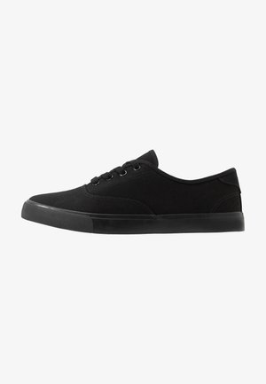 Las zapatillas negras de lona presentan un diseño de perfil bajo, punta redondeada, cordones y una suela de goma negra texturizada. Construcción simple y versátil.