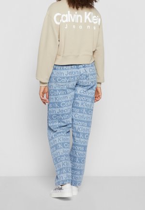 Osoba nosící béžovou mikinu Calvin Klein, světle modré džíny s opakujícím se vzorem loga Calvin Klein Jeans a bílé tenisky Calvin Klein.