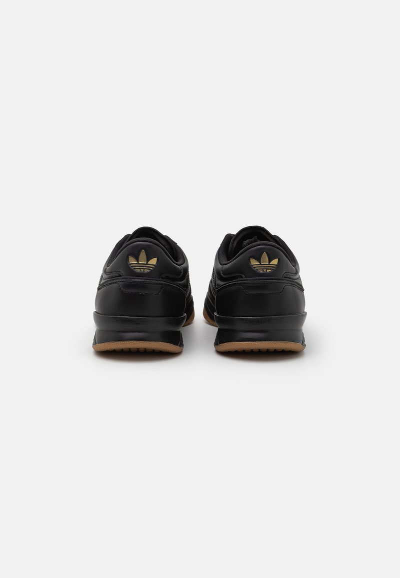 Zapatillas deportivas negras con una parte superior sintética lisa, que presentan un acento en logo dorado y una suela de goma marrón. Diseño de talón reforzado.