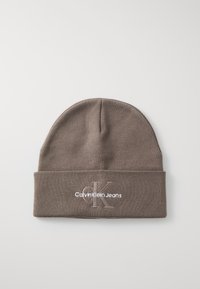Smeđa pletena kapa s manžetom. Ima bijeli izvezeni logo s natpisom "Calvin Klein Jeans" i stiliziranim "CK". Glatka tekstura.