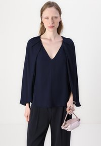 Blusa blu navy con scollatura a V profonda e maniche a mantello. Tessuto liscio, vestibilità rilassata, abbinata a una piccola borsa rosa chiaro.