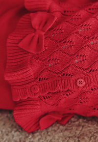 Rood gebreid kledingstuk met een gestructureerd patroon met golvende lijnen, twee knopen en decoratieve strikjes op de rand van de schouder en mouwen.