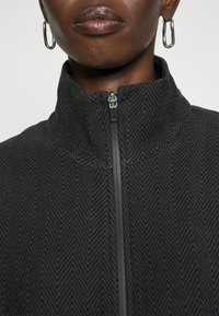 Giacca nera con zip, caratterizzata da un motivo a spina di pesce testurizzato, con colletto alto e una cerniera liscia sul davanti.