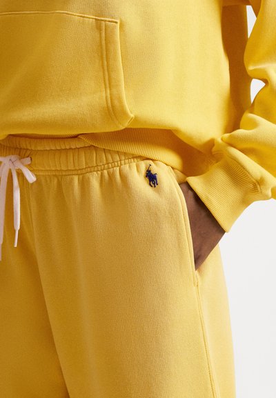 Gul bomullsgenser med hette og en frontlomme, sammen med matchende gule joggebukser med elastisk midjebånd og en blå brodert logo.