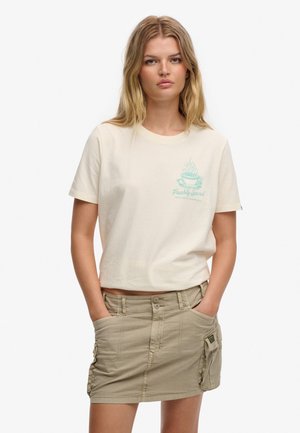 Młoda kobieta z długimi blond włosami ma na sobie kremowy t-shirt z turkusową grafiką parującej filiżanki oraz beżową spódnicę cargo, ręce w kieszeniach.