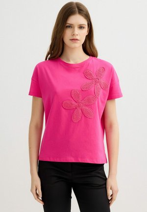 Joven mujer que lleva una camisa de manga corta de color rosa brillante con dos grandes apliques florales en relieve, combinada con pantalones negros.