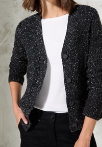 Czarny dziany kardigan z białymi nakropieniami, z dekoltem w serek, długimi rękawami i czterema dużymi czarnymi guzikami. Noszony na białej bluzce.