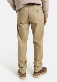 Chinos beige avec une coupe ajustée, dotés de deux poches arrière avec fermetures à bouton et d'une texture lisse, associés à des chaussures marron.