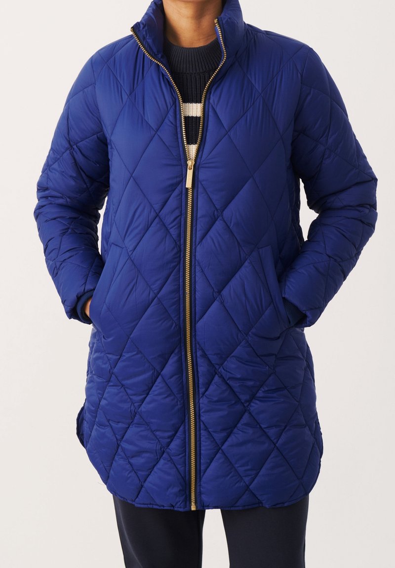 Veste longue bleu marine matelassée avec fermeture éclair dorée, portée sur un pull noir à rayures blanches, les mains dans les poches avant.