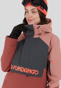 Różowa i szara wodoodporna kurtka z wysokim kołnierzem, zapinanymi na zamek kieszeniami oraz wyraźnym pomarańczowym logo na przedniej części i dolnym obrzeżu.