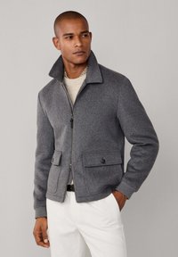 Chaqueta de lana gris con cierre de cremallera, amplio cuello y dos bolsillos delanteros. Acabado texturizado, combinada con un suéter de punto claro debajo.