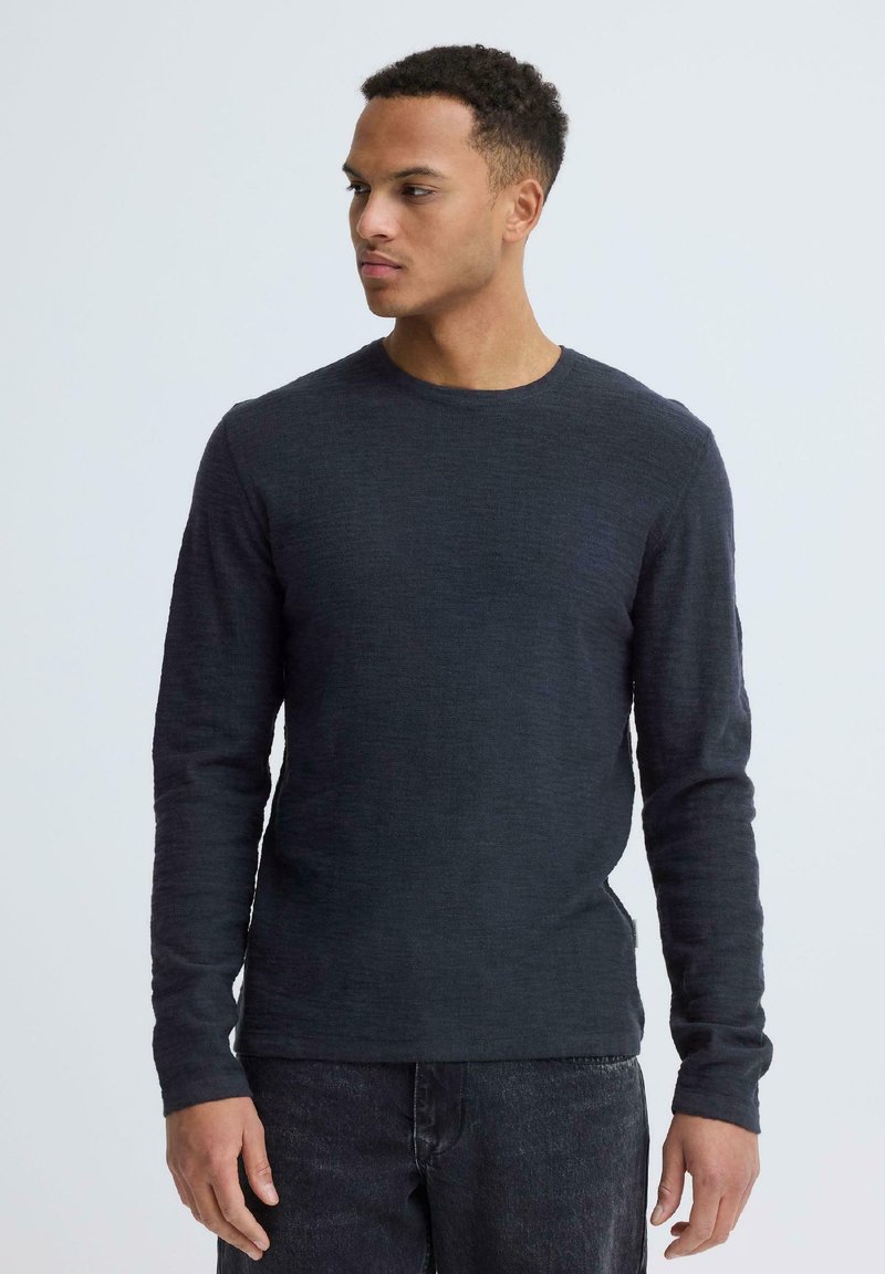 Jeune homme portant un pull à manches longues sombre et un jean noir, regardant vers sa gauche, sur un fond uni.